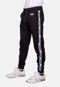 Svarta joggerbyxor med elastisk midja, dragsko och sidpaneler som visar vit Moschino-branding. Bärs med vita träningsskor.