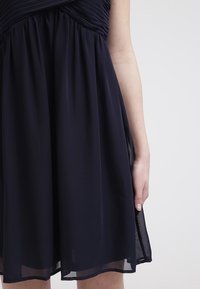 Robe plissée bleu marine foncé avec un ourlet transparent portée par une personne sur un fond clair uni, visible de la mi-cuisse au bras.