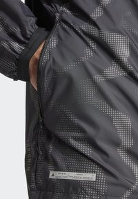 Schwarze Windjacke mit einem gepunkteten Muster. Verfügt über eine Reißverschlusstasche, elastische Bündchen und ein Etikett mit Koordinaten. Glattes, wasserabweisendes Material.