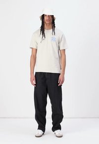 Smėlinis medvilnės marškinėliai su mėlyna grafika, derinami su juodais džinsais. Modelis dėvi baltą kibirą ir baltus sportbačius su pilkais akcentais.