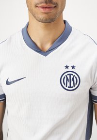 Camiseta de fútbol blanca con un patrón texturizado, cuello azul y acentos en navy. Presenta un logo circular y dos estrellas encima de él.