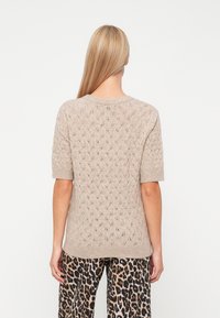 Femme aux longs cheveux blonds portant un pull en maille beige à manches courtes et un pantalon à imprimé léopard, debout de dos sur un fond blanc.