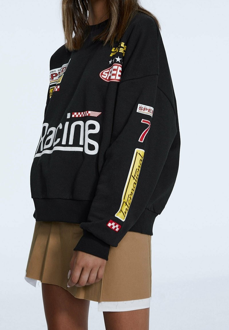 Sudadera negra con corte oversized, que presenta parches y bordados coloridos con temática de carreras. Combinada con una falda plisada color beige.