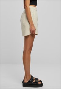 Bézs színű lenvászon rövidnadrág elasztikus derékpánttal, ívelt szegéllyel és oldalsó részletekkel, fekete papucsokkal párosítva.