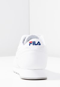 Bílé tenisky FILA s texturovaným koženým svrškem, kulatou patou a modro-červeným vyšitým logem na zadní straně. Jemné detaily šití.