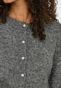Cardigan en tricot gris avec un col rond et des boutons décoratifs en perles sur le devant. Le tissu a un aspect doux et texturé.
