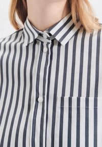 Button-up shirt with navy and white vertical stripes. Features a collar, front pocket, and textured fabric with visible stitching.