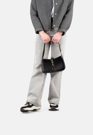 Borsa a tracolla in pelle nera con una forma curva, accessori in oro e una maniglia corta, abbinata a jeans grigi a zampa e una giacca grigia.
