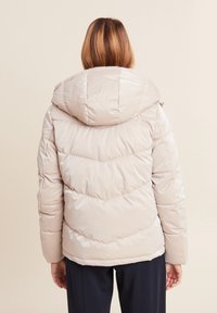 Doudoune en beige clair avec un design en chevrons, dotée d'une capuche et de détails zippés, fabriquée à partir d'un matériau lisse et brillant.