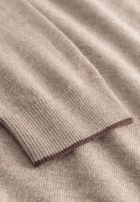 Beige Strickpullover-Detail mit einem gerippten Bündchen und einem kontrastierenden braunen Besatz, der eine weiche Textur und ein glattes Finish zeigt.