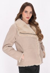 Veste en fausse fourrure beige doublée, avec un large col, une fermeture à glissière et des poches latérales. Le tissu est doux, avec une finition lisse et des coutures soignées.