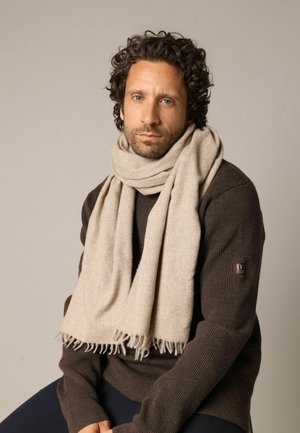 Écharpe beige en tissu doux et texturé avec des bords à franges, portée sur un pull côtelé marron foncé. Palette de couleurs simple et neutre.