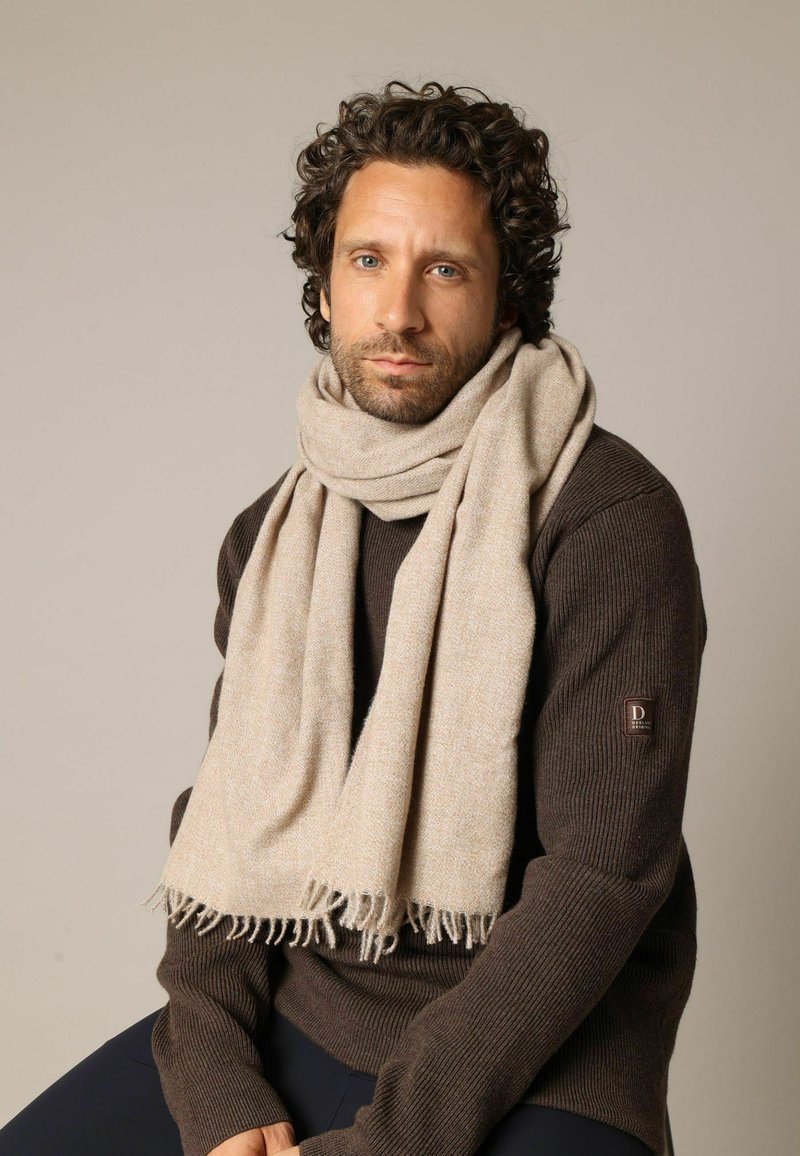 Écharpe beige en tissu doux et texturé avec des bords à franges, portée sur un pull côtelé marron foncé. Palette de couleurs simple et neutre.
