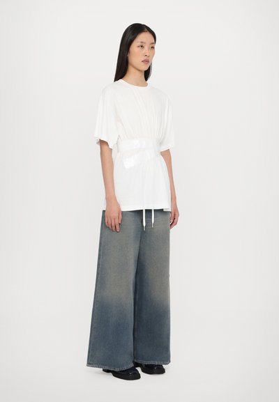 Vit skjorta med samlad textur i midjan och en glänsande bälte, i kombination med vida, urtvättade denimjeans. Modellen står framför en neutral bakgrund.