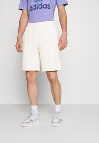 Krämfärgade shorts med elastisk midja och sidofickor, ihop med en lila skjorta med en blå logotyp. Vita sneakers visas.