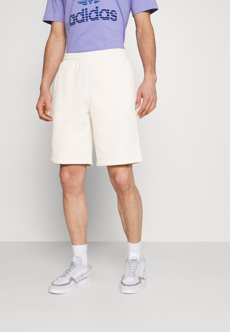 Krämfärgade shorts med elastisk midja och sidofickor, ihop med en lila skjorta med en blå logotyp. Vita sneakers visas.