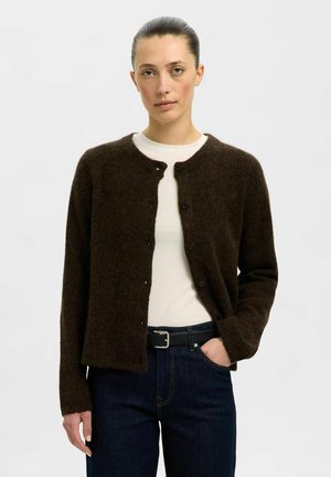 Haját hátratűzött nő, aki sötétbarna, gombos kardigánt visel fehér ing fölött, sötétkék farmerral és fekete övvel, kezét zsebében.