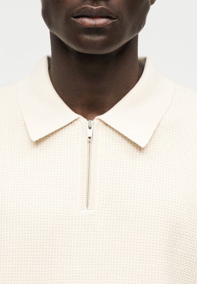 Gros plan d'une personne à la peau foncée portant un polo en maille crème texturée avec une fermeture éclair argentée au col, lèvres visibles, fond neutre.