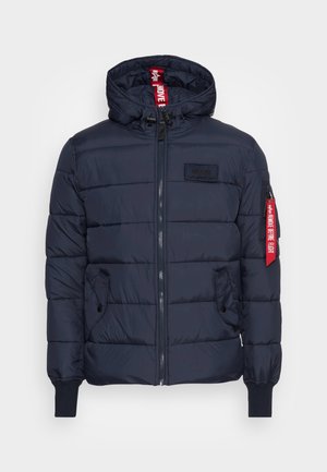 Marineblaue Steppjacke mit gestepptem Design, Reißverschluss vorne, verstellbare Kapuze, Seitentaschen, gerippte Bündchen und einem auffälligen roten Label-Akzent am Ärmel.