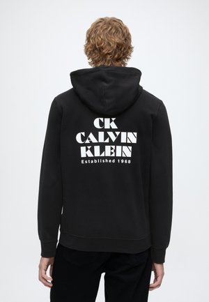 Kurde juustega inimene, kes kannab musta kapuutsiga pusa, millel on seljal suur valge tekst "CK Calvin Klein Established 1968".