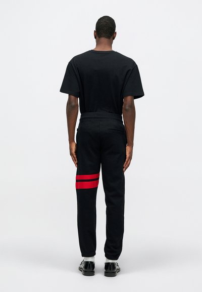 Svart kortärmad t-shirt i kombination med svarta sweatpants som har en röd randdesign på vänster ben, synliga sömmar och en avslappnad passform.