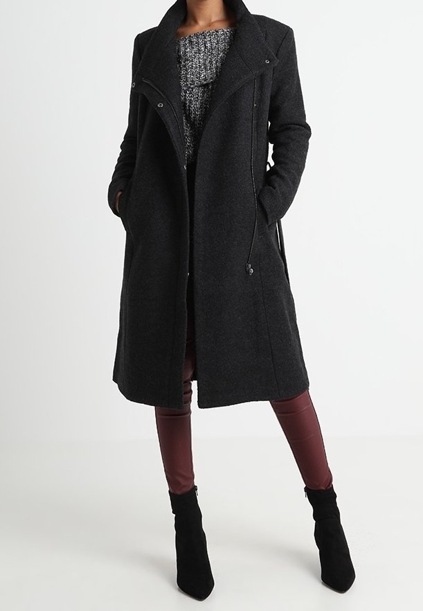 Femme portant un long manteau en laine gris foncé par-dessus un pull en maille épaisse gris, un pantalon en cuir bordeaux et des bottines noires à talons.