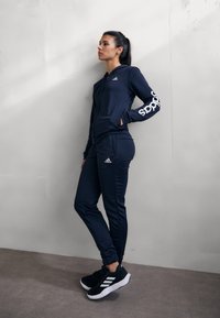 Námořnický sportovní komplet skládající se z mikiny na zip a odpovídajících kalhot, vyrobený z hladkého, elastického materiálu s bílými akcenty loga Adidas.