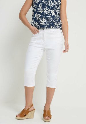 Kvinde iført hvide cropped bukser, navyblå blomster uden ærmer top og brune kilehælssandaler, der står mod en ensfarvet baggrund.