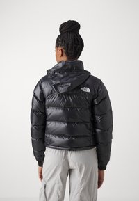 Črn pufast plašč s kvadratnim vzorcem, ki ima kapuco in logo na hrbtni strani. Gladka, sijoča tekstura z elastičnimi manšetami.