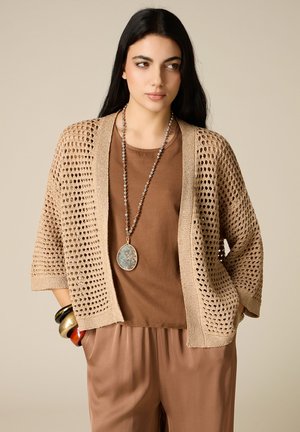 Frau mit langen dunklen Haaren trägt eine beige offene Strickjacke, braunes Oberteil, lockere Hose, große Steinanhänger-Halskette und bunte Armreifen, Hände in den Taschen.