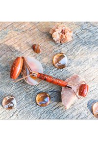 Brown crystal face roller with a rose gold metal frame on textured stone. Surrounded by agate slices and smooth, round glass stones.