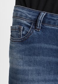 Acercamiento al área del bolsillo de unos jeans de mezclilla azul con un lazo de cinturón visible y una tela negra metida dentro de la cinturilla.