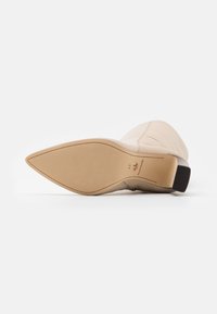 Beige klackboot, spetsig tå, syntetiskt material, präglad design på ovandelen, slät textur, platt gummisula och minimala hårdvarudetaljer.