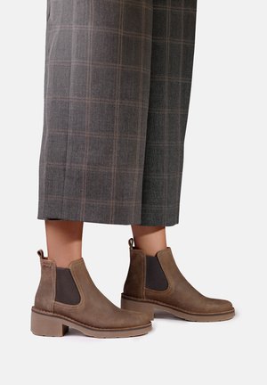 Bottines en cuir marron avec des panneaux latéraux élastiques portées avec un pantalon large gris à carreaux.