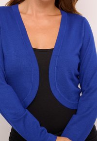Cardigan léger bleu à manches longues, à ouverture frontale, avec une texture lisse. Porté sur un haut noir, doté d'un décolleté en V.