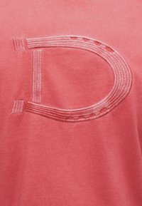 Μεγάλο ανάγλυφο και κεντημένο γράμμα "D" σε ροζ νήμα που ταιριάζει, σε φόντο από απαλό ύφασμα κοραλλί χρώματος.