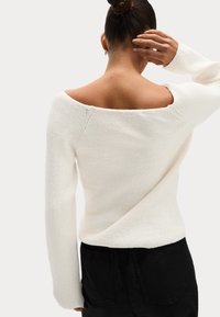 Femme avec les cheveux en chignon portant un pull en maille blanc à épaules dénudées et un pantalon noir, la main touchant l'arrière de sa tête.