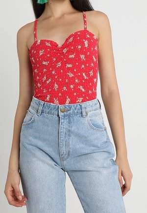 Vrouw draagt een rood met bloemen bedrukt geplooid hemdje met spaghettibandjes en lichtblauwe high-waist jeans.