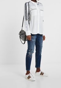 Witte blouse met gedetailleerde print op de mouwen, gecombineerd met versleten blauwe jeans en witte sneakers met gouden accenten en tekst. Zwarte schoudertas.
