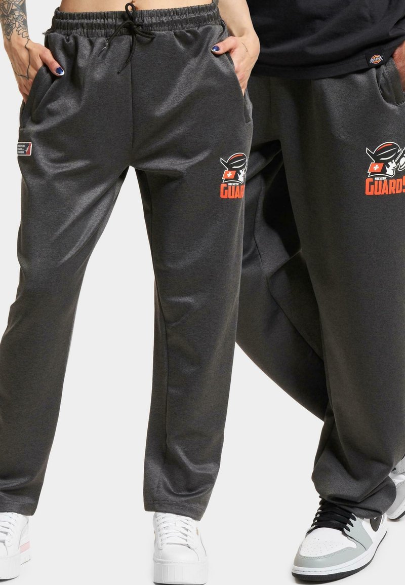 Grå sweatpants med elastisk talje og snøring, med sidelommer og et logo i orange og sort på venstre lår.