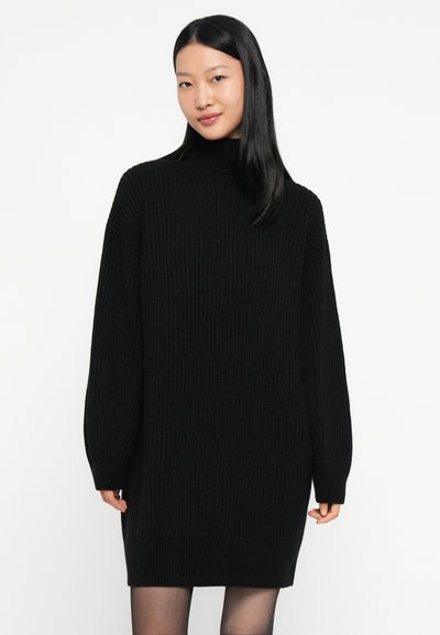 Jeune femme portant une robe pull en maille côtelée noire à col roulé et à manches longues sur fond blanc.