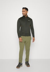 Olive green zip-up jacket with orange accents, slim fit and high collar, paired with matching olive pants and gray hiking shoes.