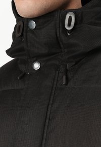 Veste noire en tissu texturé, avec un col haut et des accents de boutons en argent. Dotée d'une capuche avec cordon de serrage et d'éléments de design épurés.