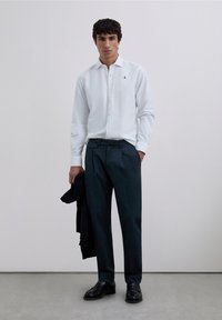 Cămașă albă cu mâneci lungi și nasturi, pantaloni eleganți verzi închis, pantofi negri. Modelul ține un pulover bleumarin. Design simplu cu branding subtil.