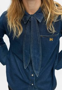 Camicia in denim blu scuro, con colletto a punta e design a papillon, dotata di taschino sul petto. Cuciture chiare e piccolo dettaglio con logo.