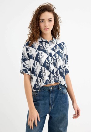 Blusa corta de mangas cortas con un patrón de langostas en azul marino y blanco sobre una tela ligera. Combinada con jeans azul oscuro de tiro alto.