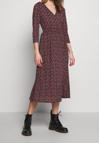 Blumige Wickelkleid in Dunkelrot mit Dreiviertelärmeln, V-Ausschnitt, elastischem Taillengummi und fließender Midi-Länge. Getragen mit schwarzen Schnürstiefeln.