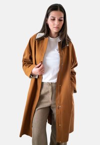 Jonge vrouw die een open bruine jas over een wit shirt en beige broek aanpast, staande tegen een lichte achtergrond.