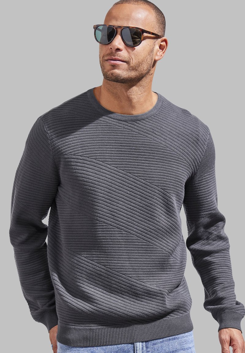Grauer strukturierten Pullover mit schräger Rippung, rundem Halsauschnitt und gerippten Bündchen. Kombiniert mit dunkler Sonnenbrille und blauen Jeans vor einem neutralen Hintergrund.