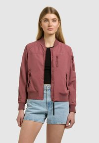 Femme portant une veste bomber mauve sur un haut noir, associée à un short en denim bleu clair à taille haute, debout devant un fond uni.